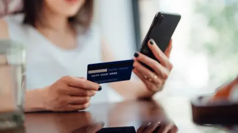 Getty Images A woman is holding a credit card or debit card and doing online shopping on a smartphone. 