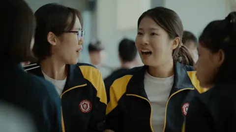 Barunson E&A Four high school girls sit together in a cafeteria. They are wearing matching black-and-yellow athletic-school school jackets. Joo-in's friend, left, is turned slightly to the side wearing glasses, while Joo-in faces forward with her mouth open mid-expression