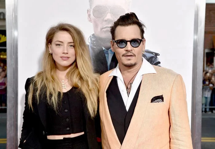 Amber Heard and Johnny Depp stand on a red carpet. One wears a black outfit, and the other is in a beige suit with a white shirt and sunglasses