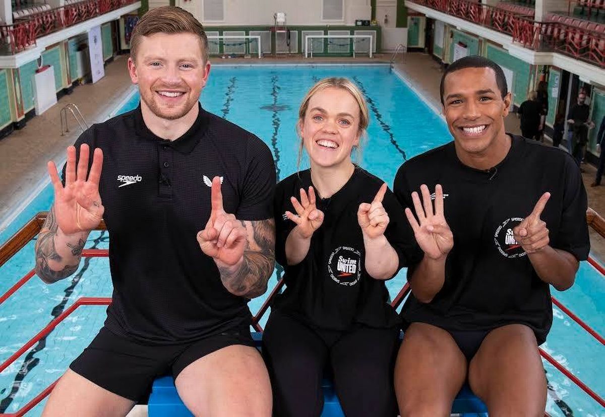Three individuals dressed in black swimming suits are sitting on the edge of an indoor swimming pool, each displaying a unique hand gesture. The pool behind them is filled with clear blue water.