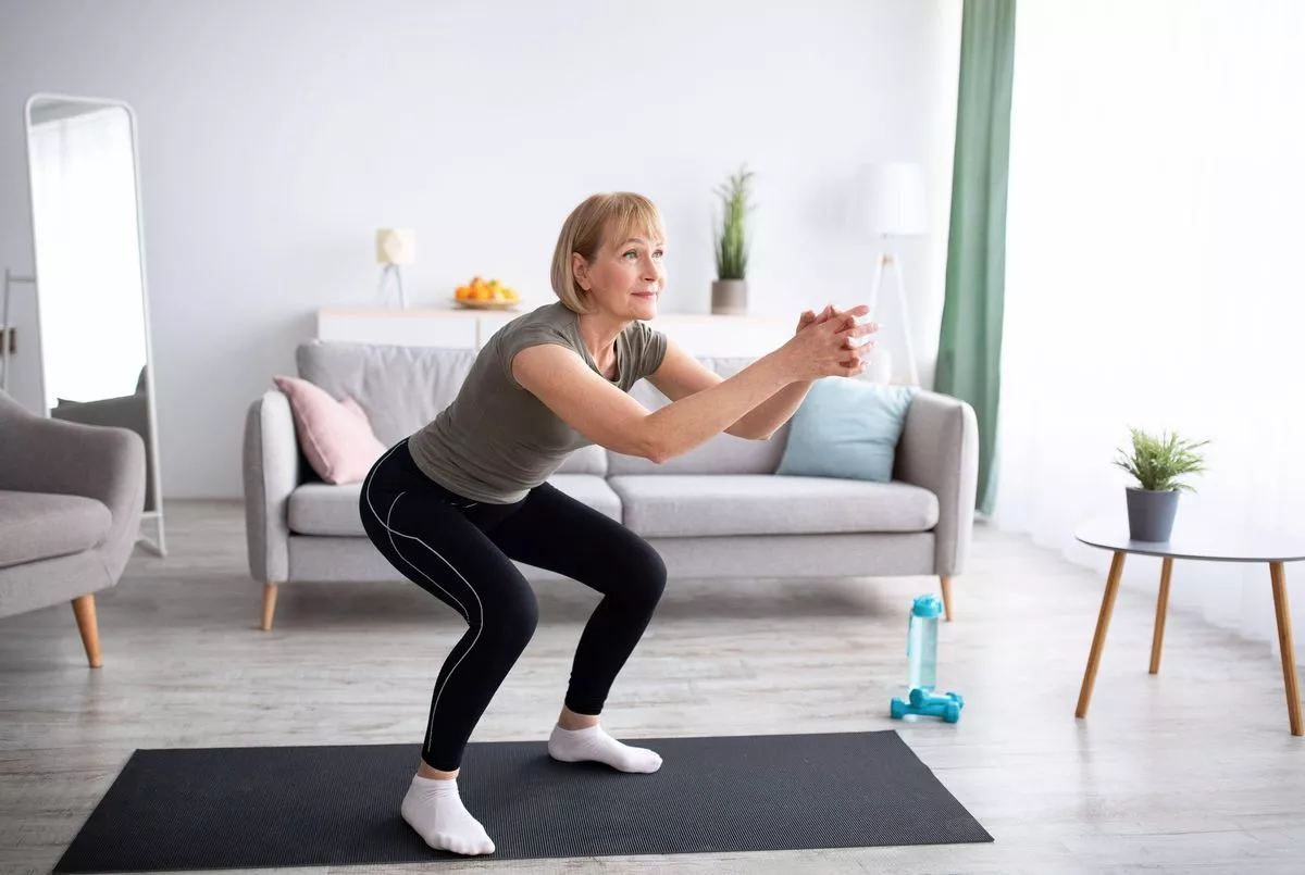 An individual is performing a workout exercise in a well-lit room with modern furnishings. The individual is dressed in a gray t-shirt, black leggings, and white sneakers, and is standing on a black mat on a polished floor. In the background, there is a white sofa with decorative pillows, a side table with a small potted plant, and a window with curtains. The setting suggests a comfortable and modern living space.