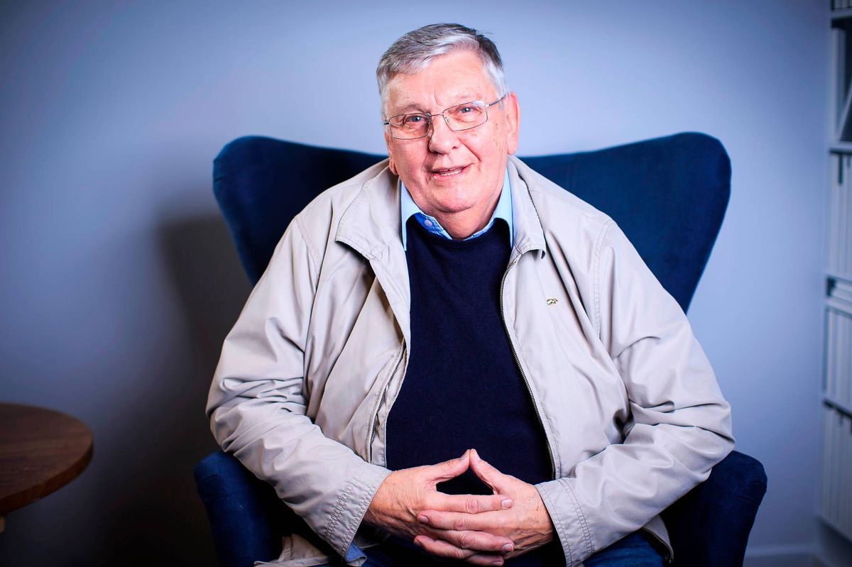 An older individual, clad in a light-colored jacket and dark top, is seated on a blue chair with a contemplative expression and folded hands. The background features a light blue wall and a glimpse of a wooden table.