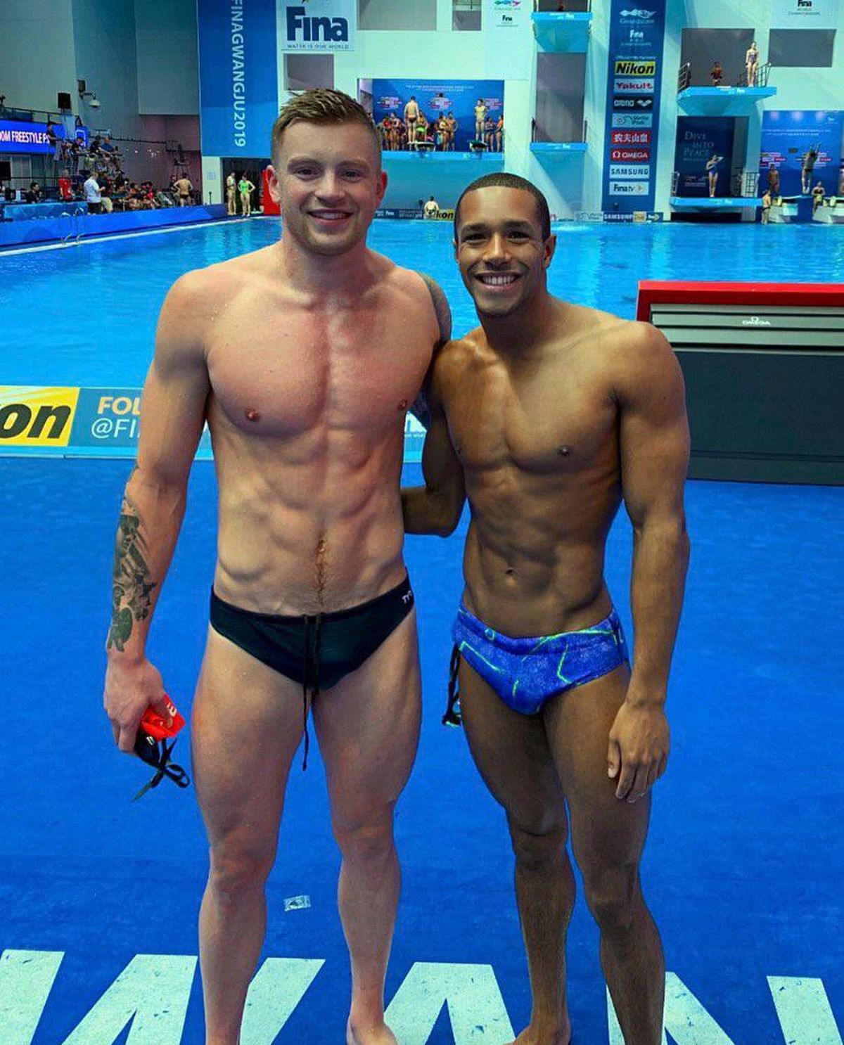 Two male athletes are standing side by side, smiling and posing for a photograph in an indoor swimming pool, with a blue water surface and a red diving board visible in the background.
