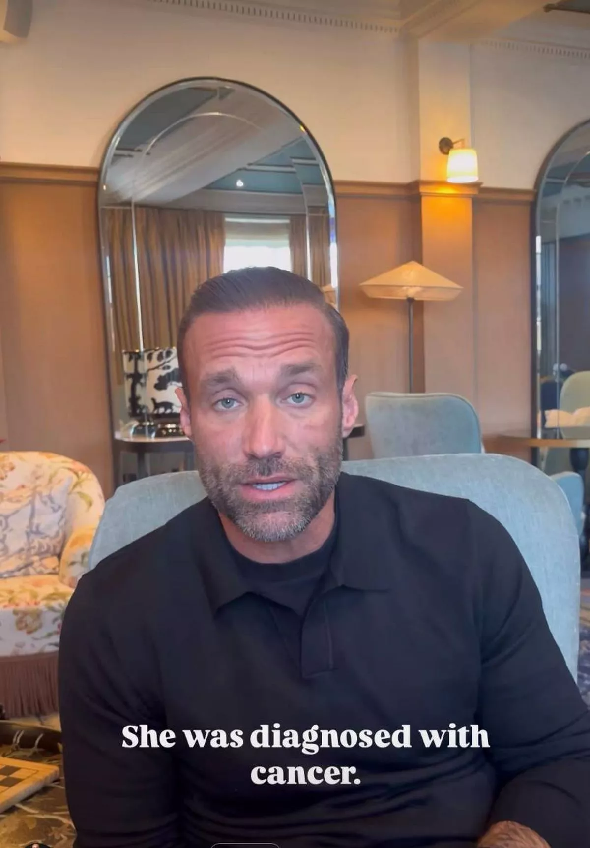An adult male, dressed in a black shirt, seated on a blue chair within an indoor setting. The individual appears to be engaged in a conversation, with various furnishings and decorative elements visible in the background.