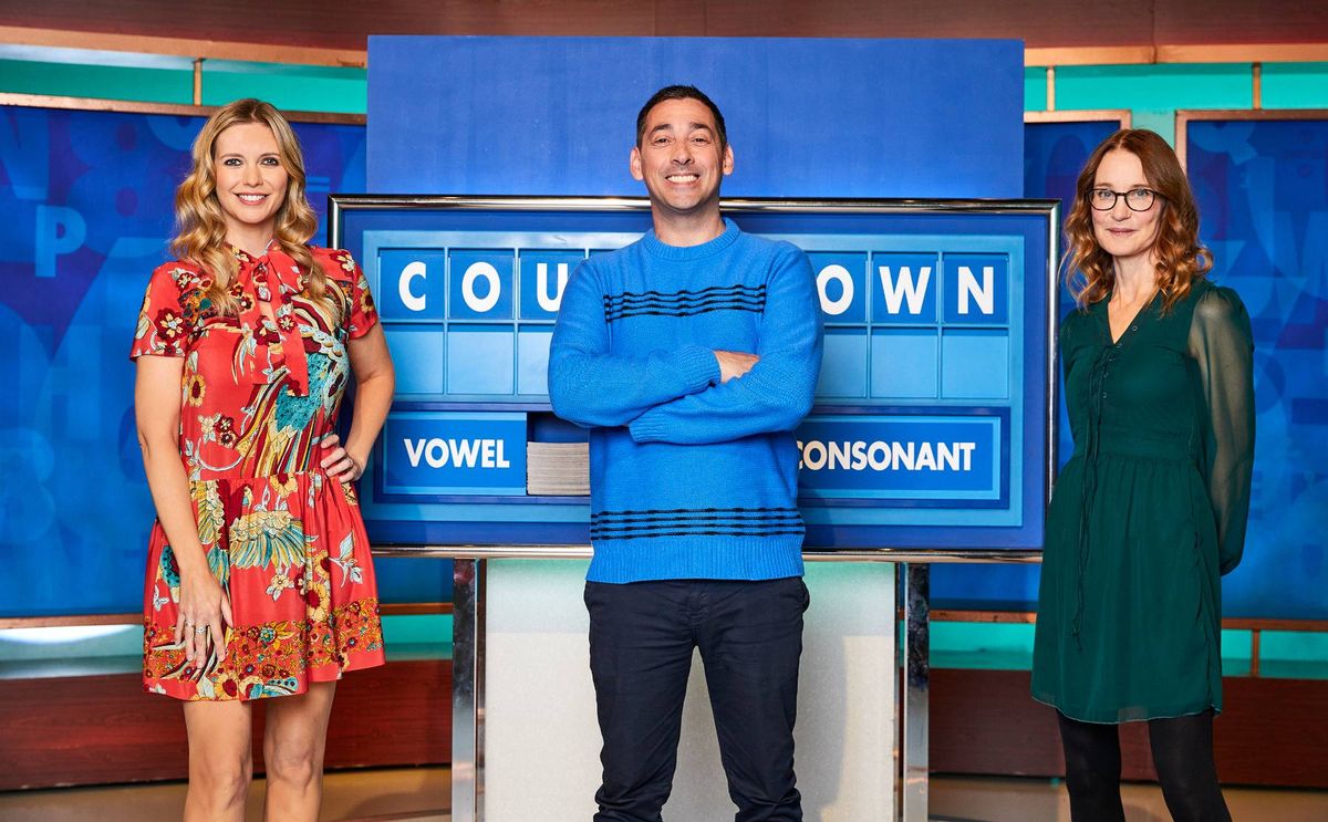 Three individuals are standing together in front of a blue backdrop that features the letters "COU OWN" prominently displayed. The central figure is a man dressed in a blue sweater, standing with his arms crossed, while the individuals on either side of him are women, one wearing a floral dress and the other a green top.