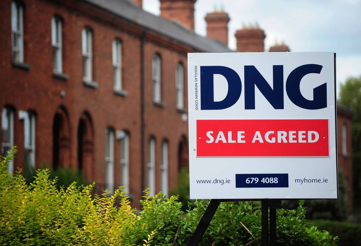 A white sign with blue and red text stating "DNG SALE AGREED" stands in front of a red brick building with several windows.