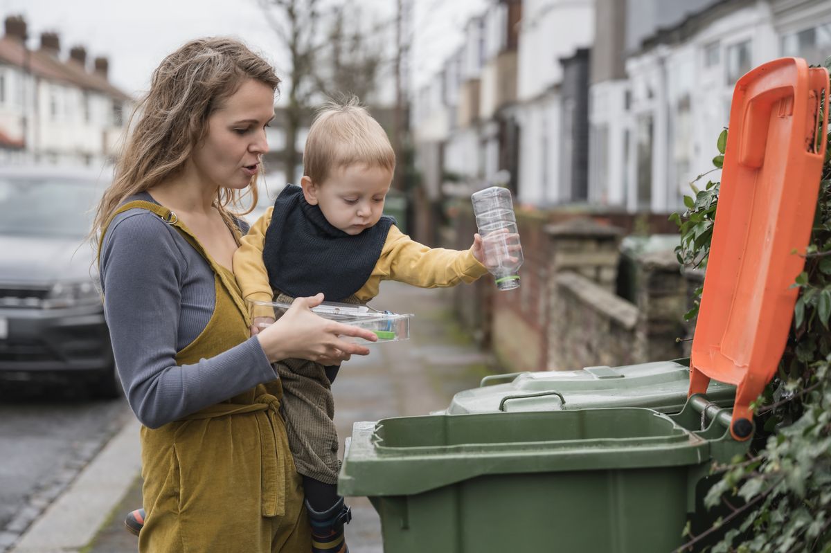 Partial side view of longhaired Caucasian woman in casual clothing holding 2 year old son in her arms as he puts plastic containers in recycling bin.
