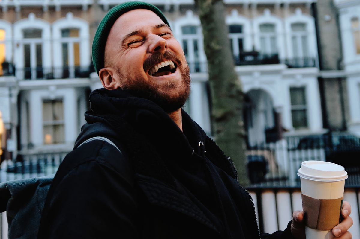 A man with a green hat is smiling and holding a coffee cup. The image has a cheerful and lighthearted mood