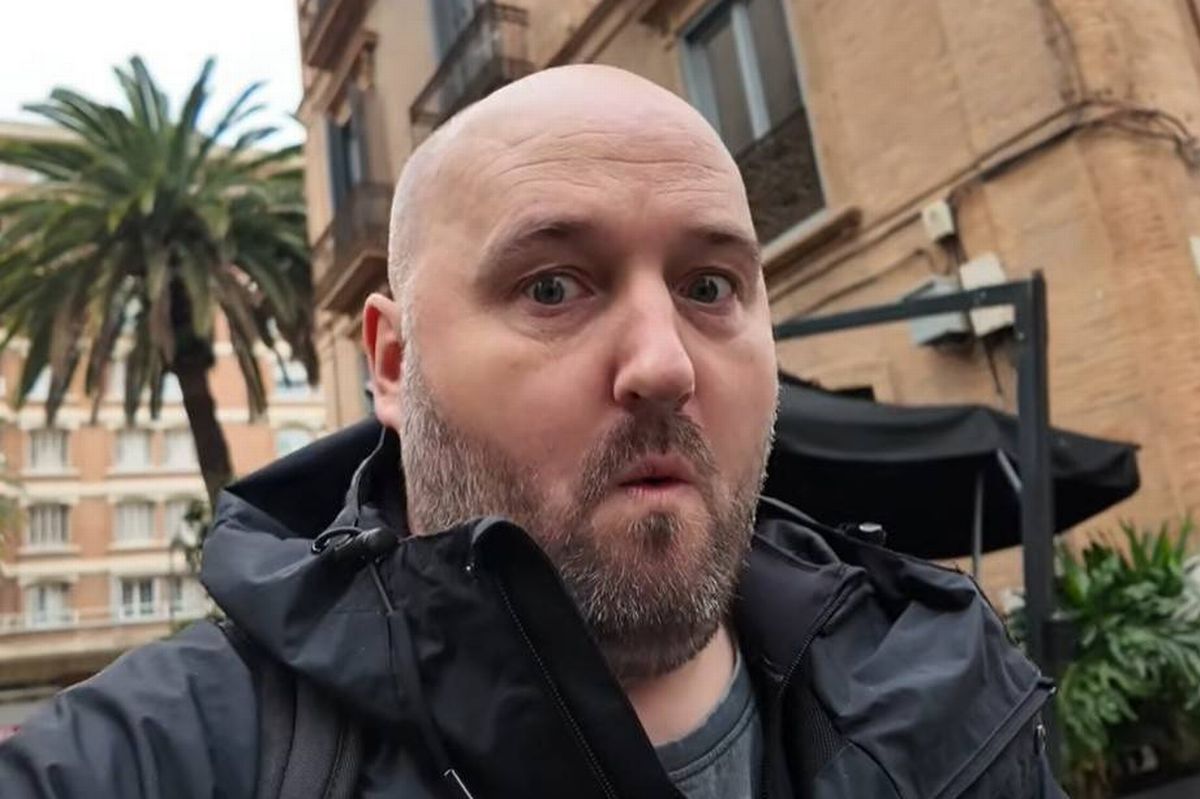 An individual with a bald head and a beard is standing outside in front of a building. He is dressed in a black jacket and appears to be taking a selfie. Behind him, there is a tree and a partially visible black umbrella. The building in the background has several windows.