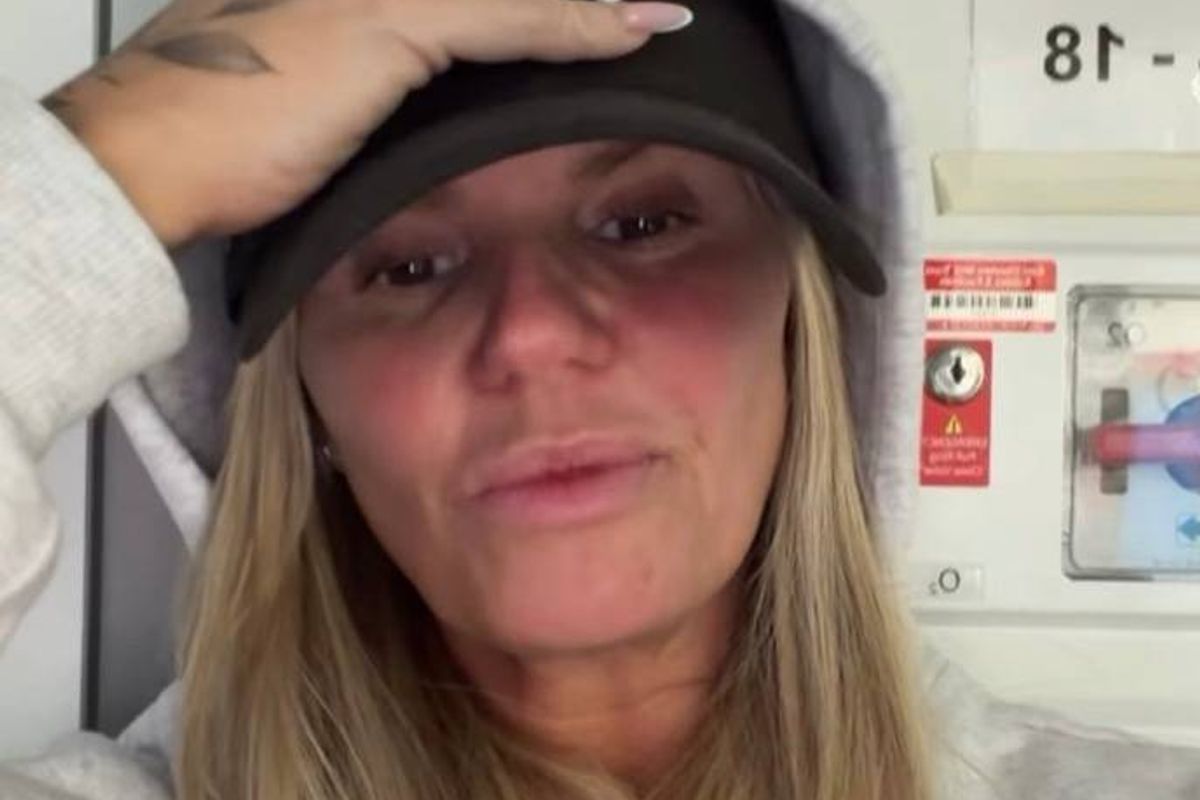 An individual with long blonde hair wearing a black cap and a white long-sleeved shirt, seemingly engaged in a video call. The background features a partially visible wall adorned with various signs and notices, one of which includes the number 81.