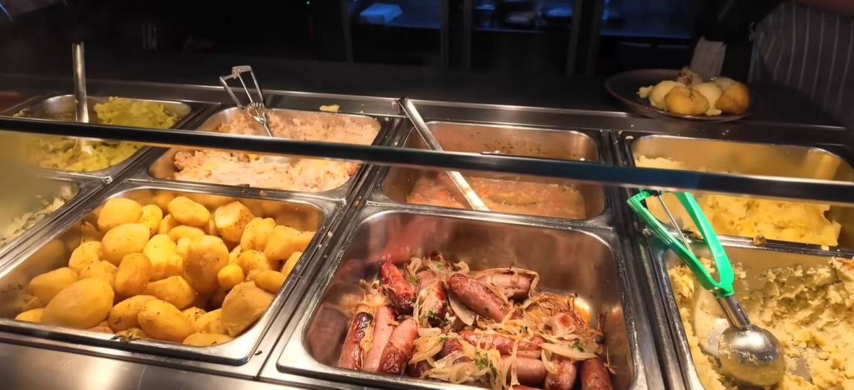 An image displaying a buffet table with various food items in stainless steel containers, including a dish of noodles and meat, accompanied by utensils for serving.
