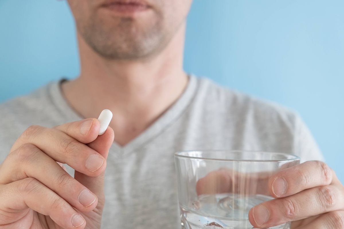 Man taking white pill of statin medicine to treat high cholesterol with glass of water on blue background. Taking medicine, health care, pharmacy and treatment concept. Selective focus