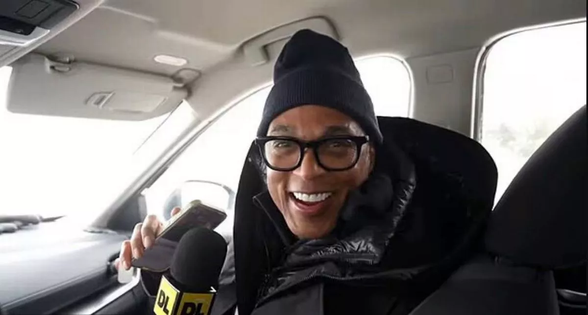 An individual seated inside a vehicle, wearing a black beanie and glasses, holding a microphone and smiling.