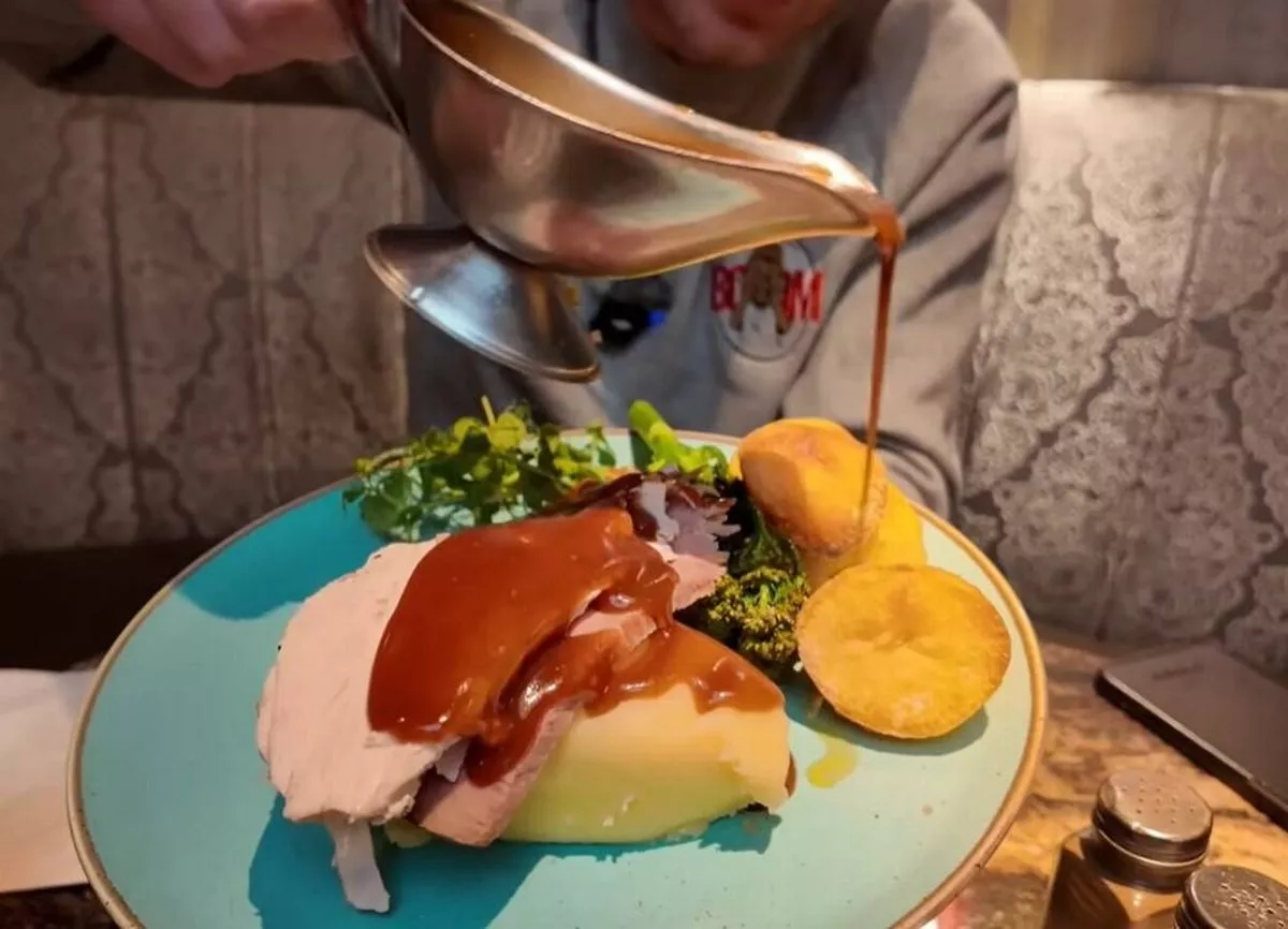 A person is pouring a sauce over a plate of food. The plate includes a slice of meat, a potato, and a salad. The food is arranged on a blue plate with a decorative border.