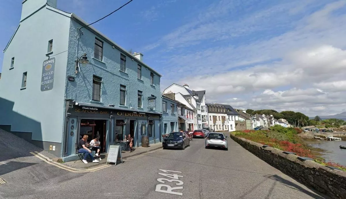 An urban street scene featuring a multi-story building on the left, with several windows and a person standing near the entrance. Vehicles are driving on the road, and the street is bordered by a sidewalk and a grassy area. The sky is partly cloudy.