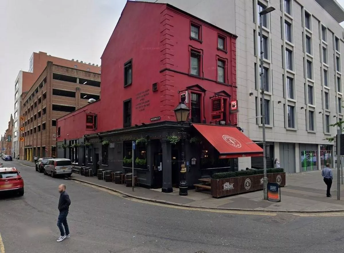 Urban street scene featuring a red building with multiple windows and an awning, situated between a modern white building and a busy road with vehicles and pedestrians.