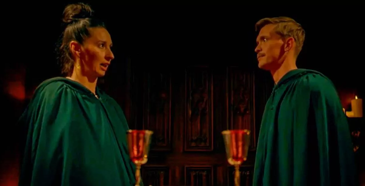 Two individuals dressed in green robes are standing close to each other, facing in the same direction, with a dimly lit background featuring a structure and candles.