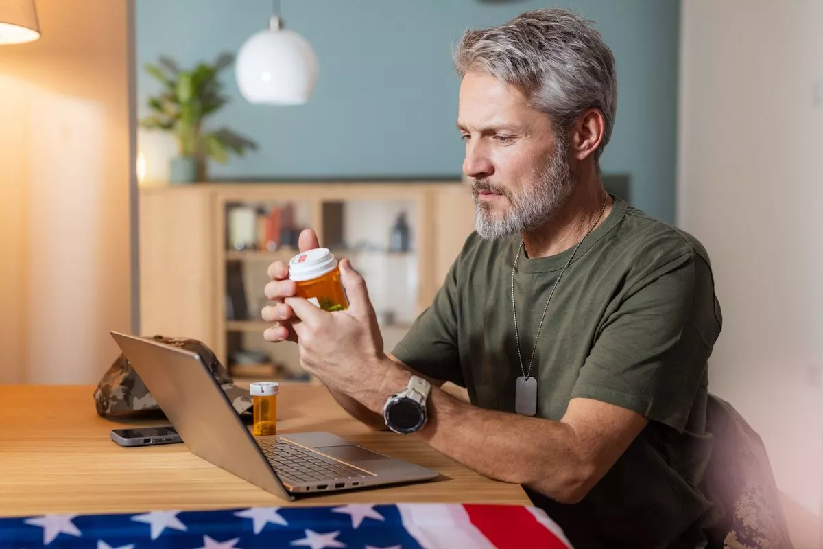 Mature white veteran man with gray hair and beard wearing casual clothes checks prescription medication bottle at home with laptop and military cap on the table under warm indoor lighting