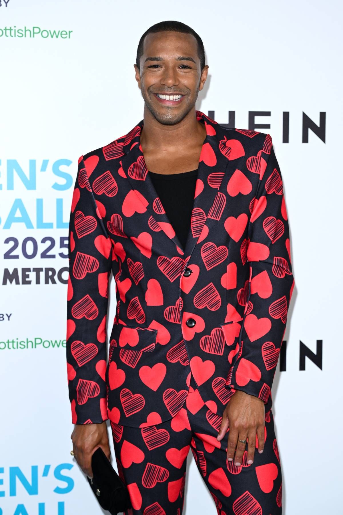An individual dressed in a vibrant red and black patterned blazer with a black inner shirt, standing in front of a backdrop featuring the word "N'S" and "EIN," smiling at the camera.