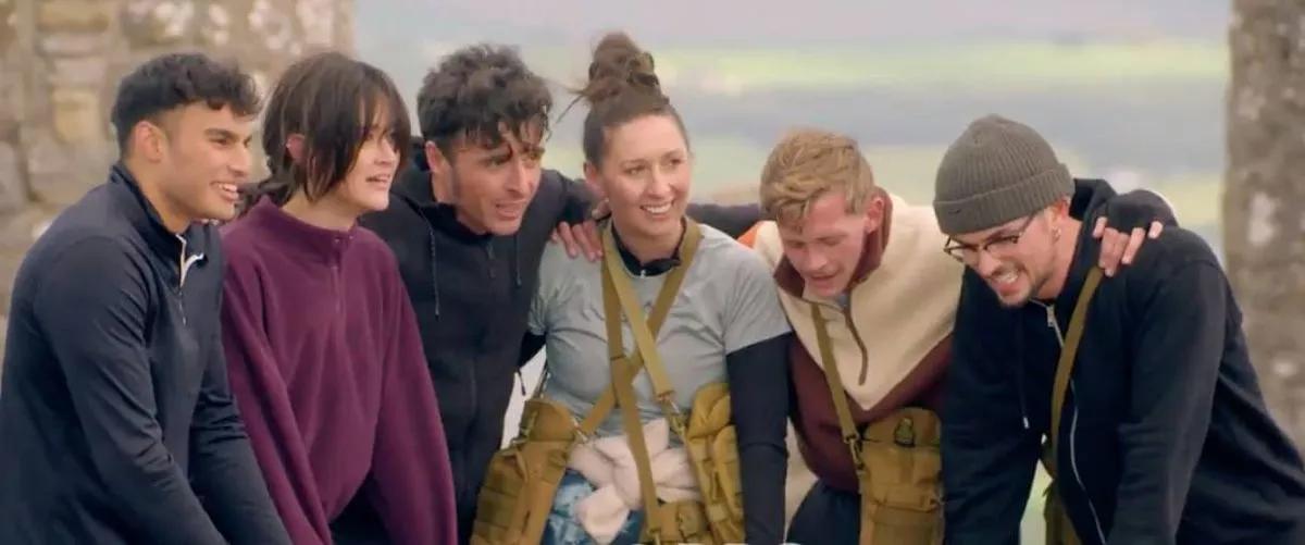 A group of individuals standing close together, smiling and posing for a photograph in an outdoor setting. The individuals are dressed in various colors, with one individual wearing a maroon top and another in a gray shirt. The backdrop suggests a scenic location, potentially near a body of water.