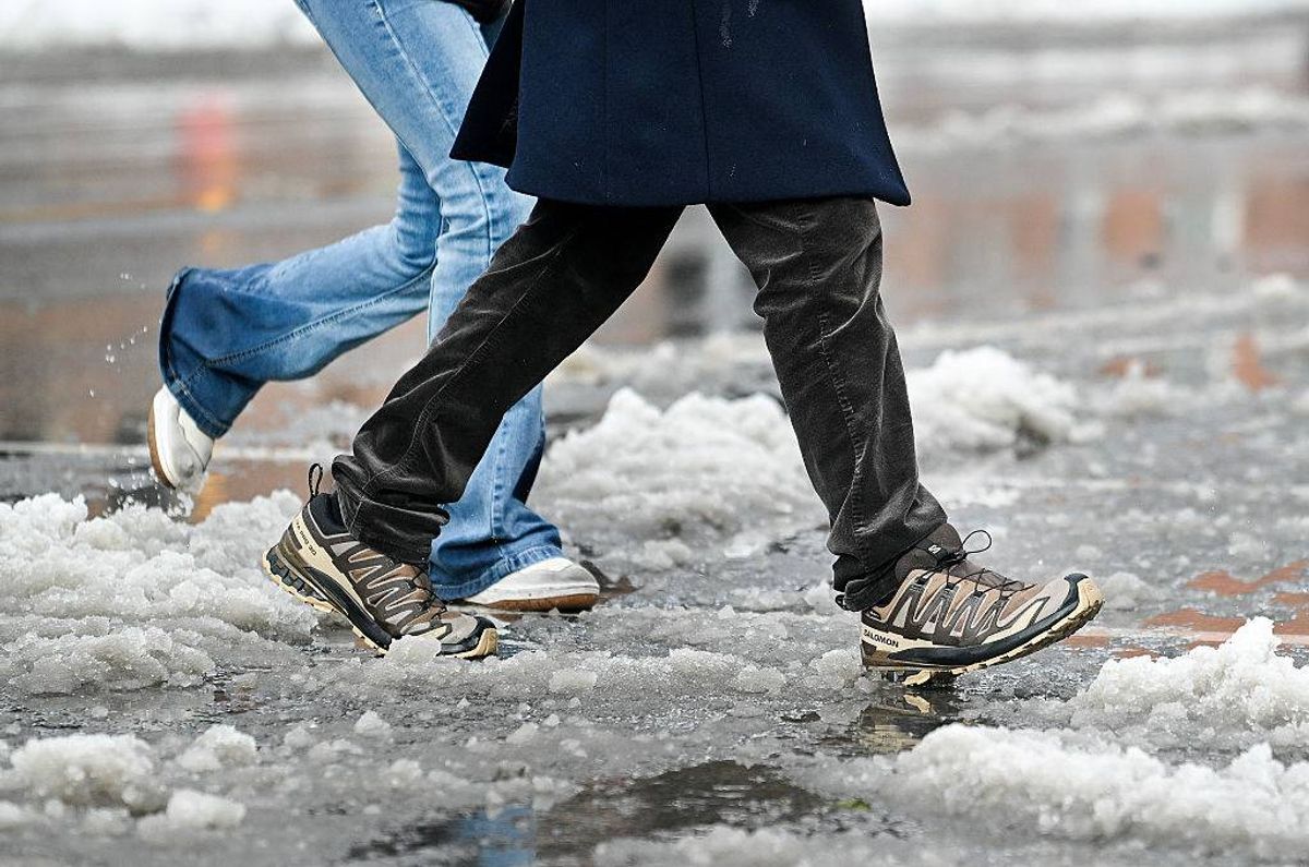 Two individuals are walking on a street covered in snow, their footwear and lower garments visibly affected by the wintry conditions. The scene indicates recent snowfall with visible footprints and some melting in areas.