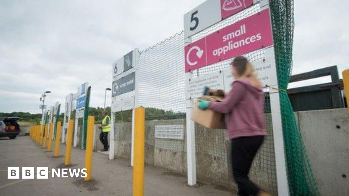 A woman carries a box as she crossed the car park of a recycling centre. A worker in a yellow high vis vest is seen entering the car park as well. The woman's figure is blurred. She is passing by a red sign that reads "Small appliances". A car with an open boot. It is a cloudy day.