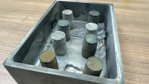A metal box, containing cylindrical metal magnets, on a wooden surface.
