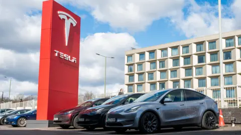Getty Images Tesla cars sit parked outside at a dealership in London. They are parked beside a red Tesla sign. 