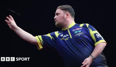 A side-on view of Luke Little as he throws a dart. He has dark hair, brushed  forward, and a beard. His shirt is blue with yellow trim.