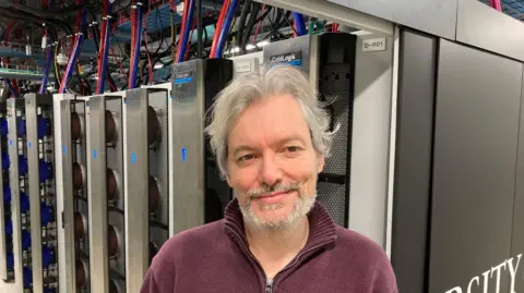 David Webster/BBC Dr Paul Calleja stands in front of one of the rows of servers. There are about eight tall silver units behind him, each with a thick blue and red cable connecting the units to the ceiling. The units have a sign on saying ColdLogik.