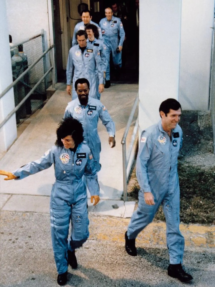 Seven people wearing blue overalls and boots walk out of a building into daylight, smiling and waving.