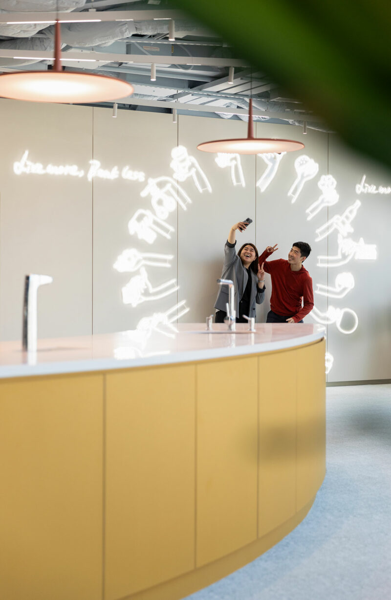 Two people stand together in an office space, taking a selfie in front of a curved counter and an illuminated wall with white neon designs.