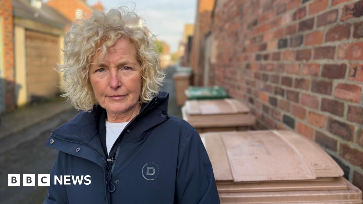 A woman with blonde curly hair is wearing a dark blue coat. She is standing in a alleyway beside brown wheelie bins and a high red-brick wall.