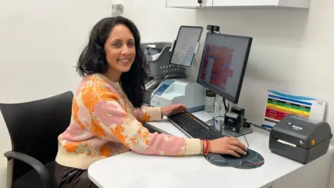 Matt Knight/BBC Hema Patel sits at a white desk on a black office chair. She has a computer monitor, keyboard and mouse in front of her and is resting her hand on the mouse. There are also several other peripheral devices on the desk. She is wearing a light coloured jumper with pink and orange patterning. She has black hair and is smiling at the camera.