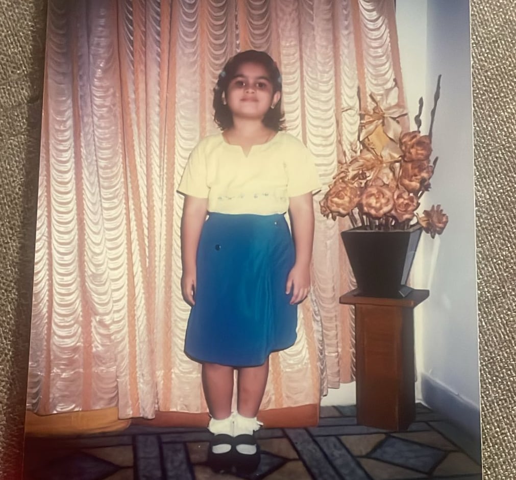 A girl wearing a yellow top and blue skirt stands very straight while posing for a photo. 