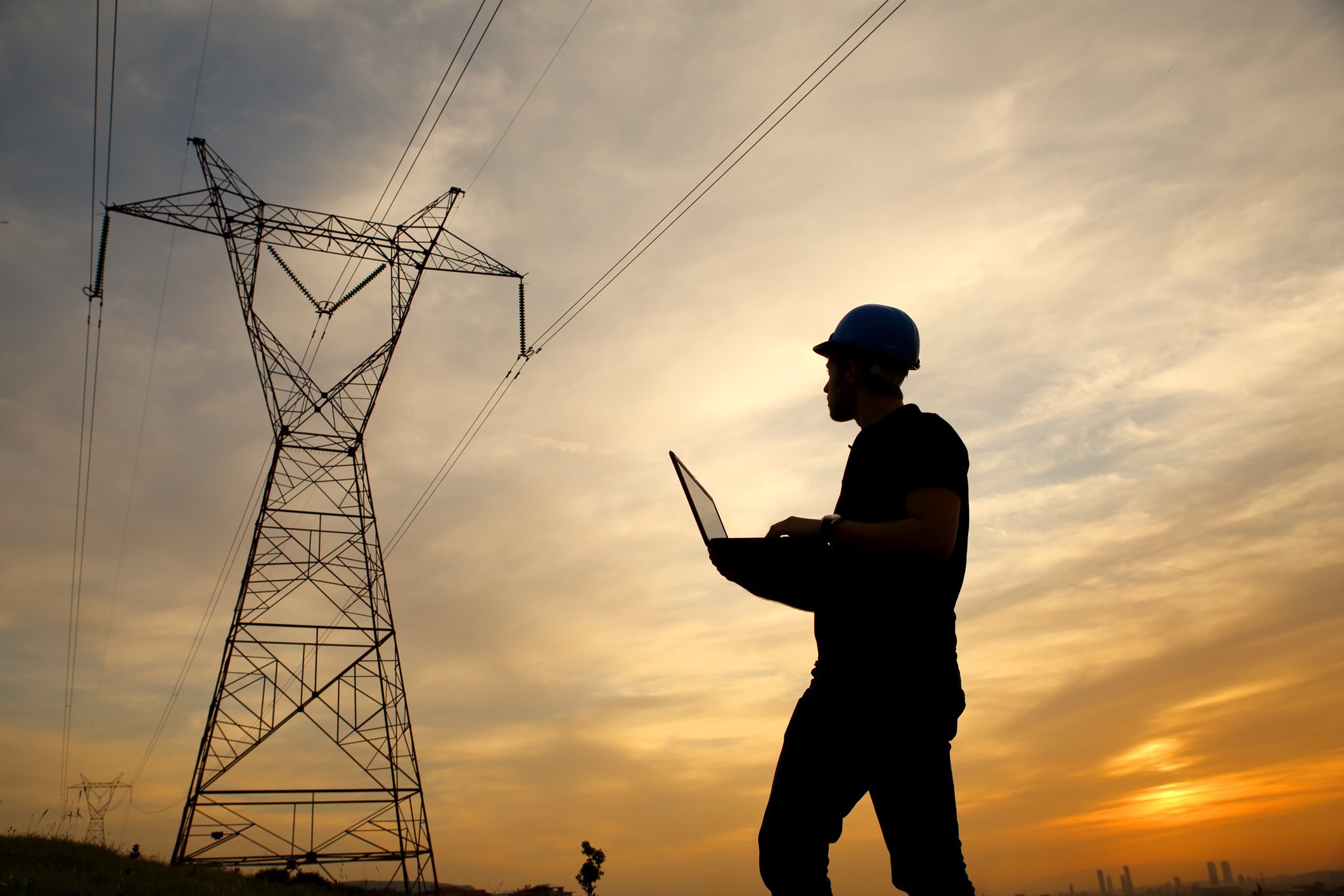 A person wearing a hardhat and holding a laptop near a power line.