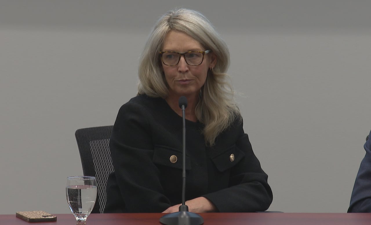 A woman with long hair sits in front of a microphone at a table.