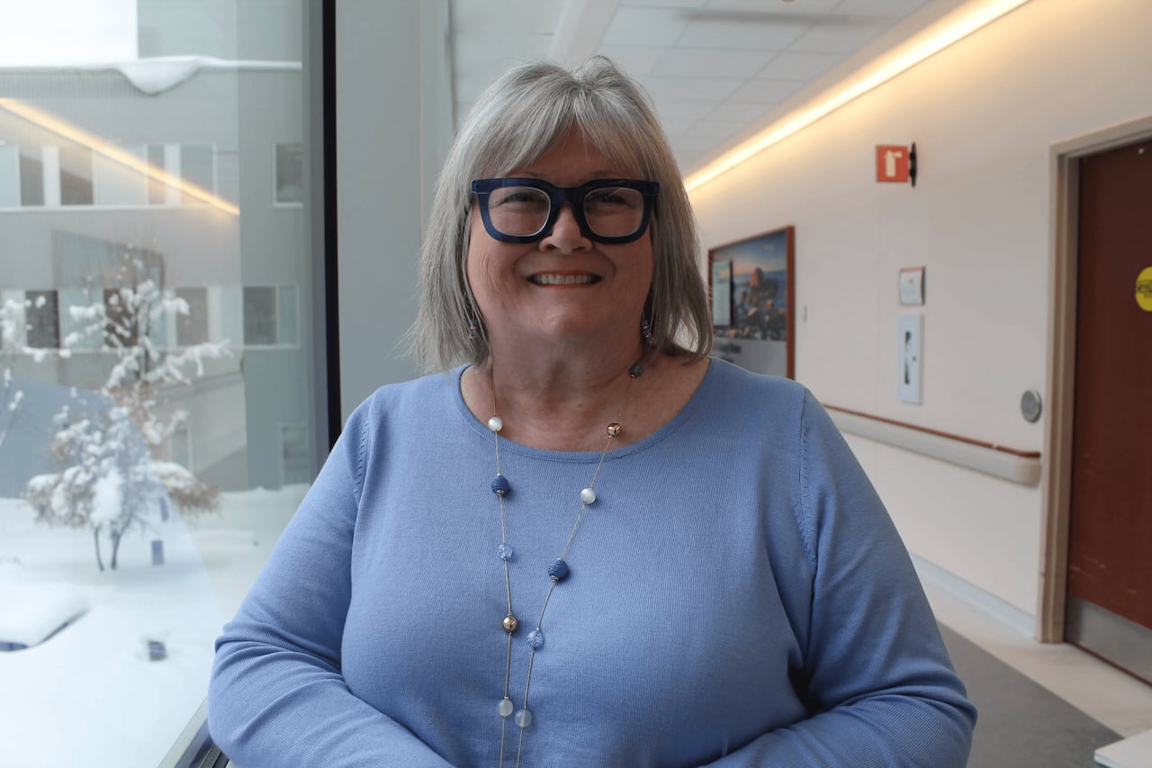 A smiling woman with a grey bob and blue framed glasses with a light blue sweater stands in front of a window