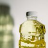 A close-up photograph of canola oil in a plastic bottle. The shadow of the bottle is cast to the left of the bottle on the white wall behind the bottle of seed oil.