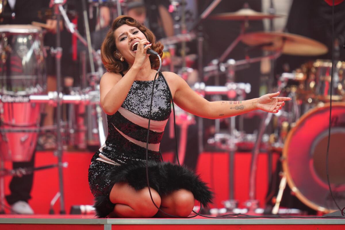 Raye in a black dress singing into a microphone while she kneels on a stage. 
