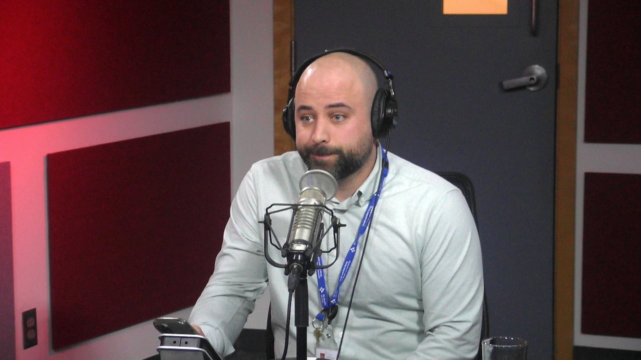 A man wearing a white shirt sits behind a microphone in a radio studio.
