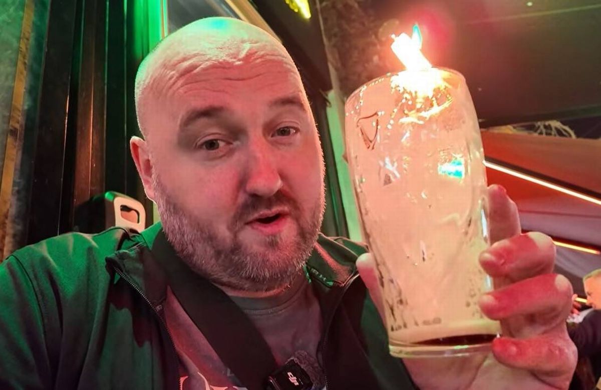 An individual dressed in a green shirt is holding a lit candle inside a glass container, positioned in a dimly lit setting.