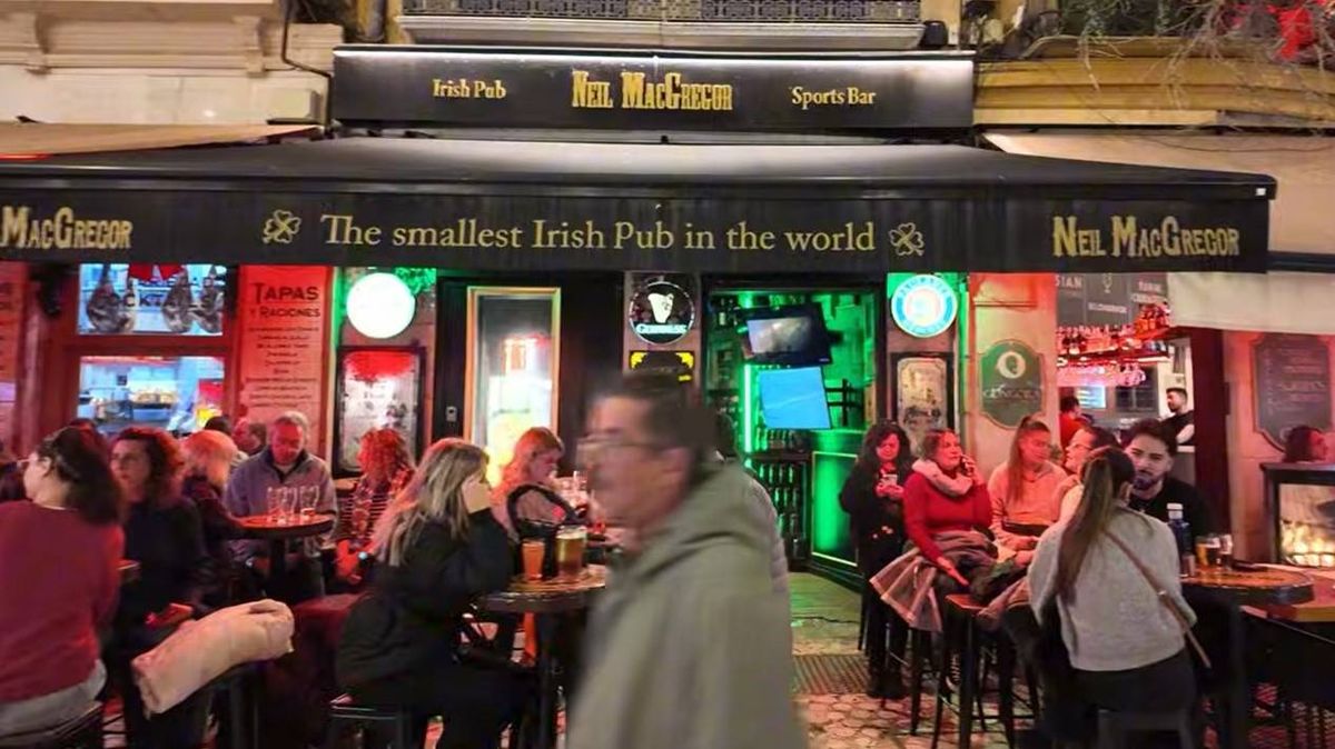 A bustling establishment with a black awning displays a neon sign featuring the phrase "The Best Irish Pub in the World." Numerous patrons are gathered both inside and outside, with some seated at tables, while a man walks past the entrance. The atmosphere is lively, with vibrant lights and signs adorning the front of the venue.