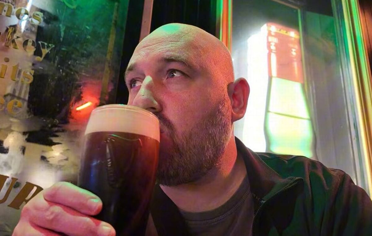 An individual with a beard is holding and drinking a glass of dark beer in an illuminated bar setting.