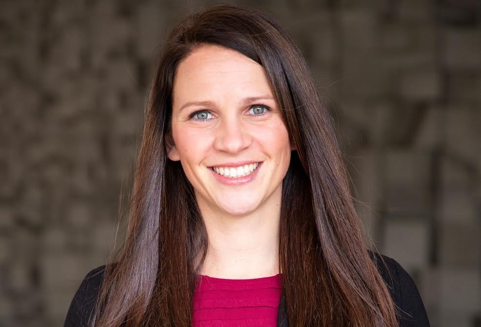 A woman with long brown hair, a black blazer and a pink shirt smiles at the camera, with a generic background. 