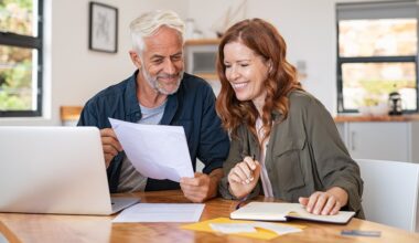 Mature couple sitting at a table, holding papers and working on their finances on a laptop in their house