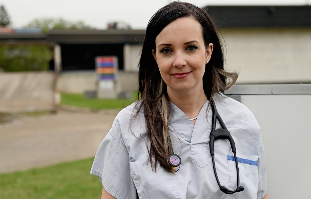 A woman with long hair wears doctor scrubs and has a stethoscope around her neck. She stands outside.