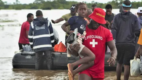EPA/Shutterstock A man in a red T-shirt and hat with the word 'Lifeguard' on helps a girl through flood waters in southern Mozambique. Behind them is a rubber speed boat with men around it.