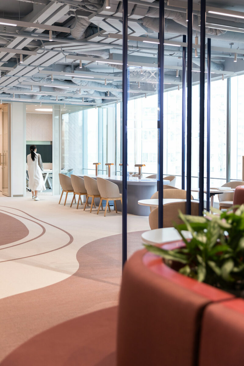 Modern office space with neutral-tone seating, large windows, and exposed ceilings; a person in white walks toward a meeting room in the background.