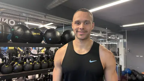 A picture of a man smiling. He is wearing a vest and standing in front of a rack of medicine balls and kettlebells.